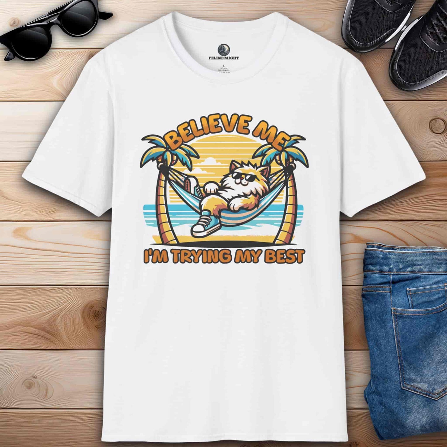 A white t-shirt featuring a cartoon cat relaxing in a hammock with palm trees and the text 'Believe Me I'm Trying My Best'.