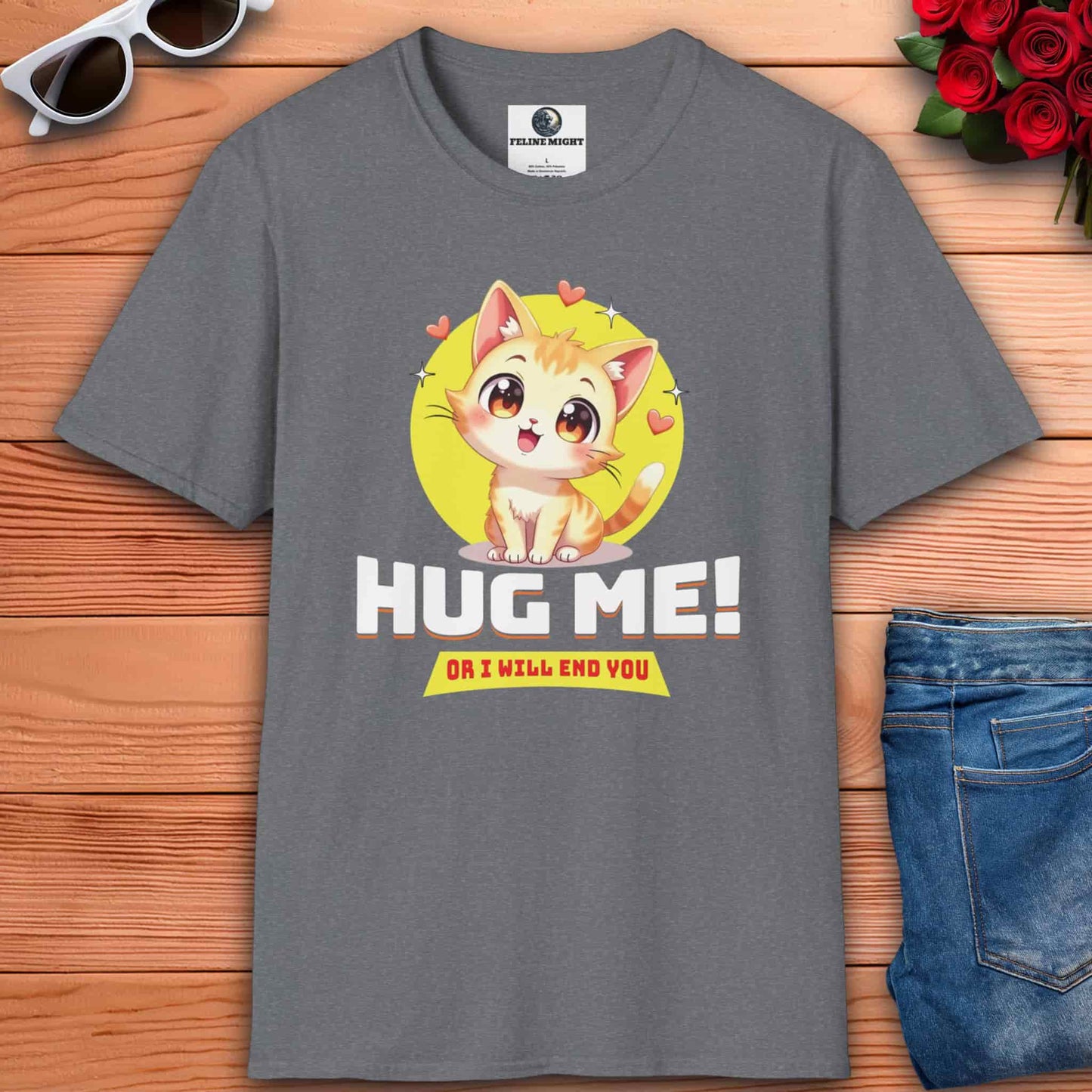 Athletic heather grey t-shirt with a cartoon cat and text: "Hug Me! Or I will end you." Surrounded by hearts, laid on a wooden surface next to sunglasses, a red rose bouquet, and blue jeans.