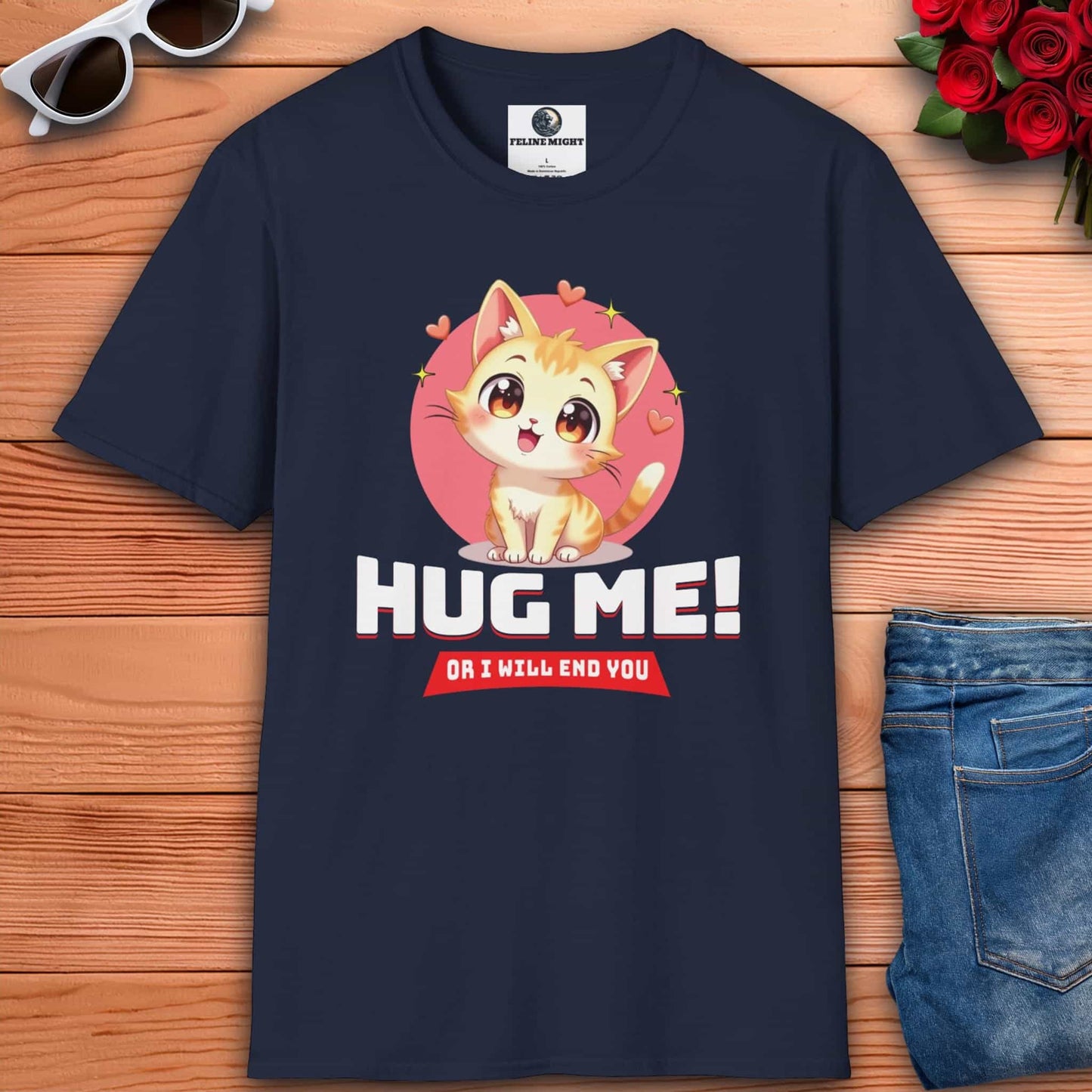 Navy blue t-shirt with a cartoon cat and text: "Hug Me! Or I will end you." Surrounded by hearts, laid on a wooden surface next to sunglasses, a red rose bouquet, and blue jeans.