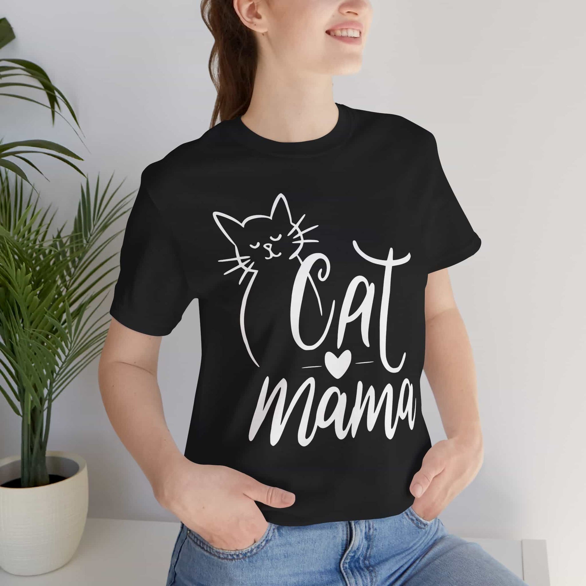 A woman wearing a black Cute Cat Mama T-shirt with 'Cat Mama' design, standing indoors next to a plant.