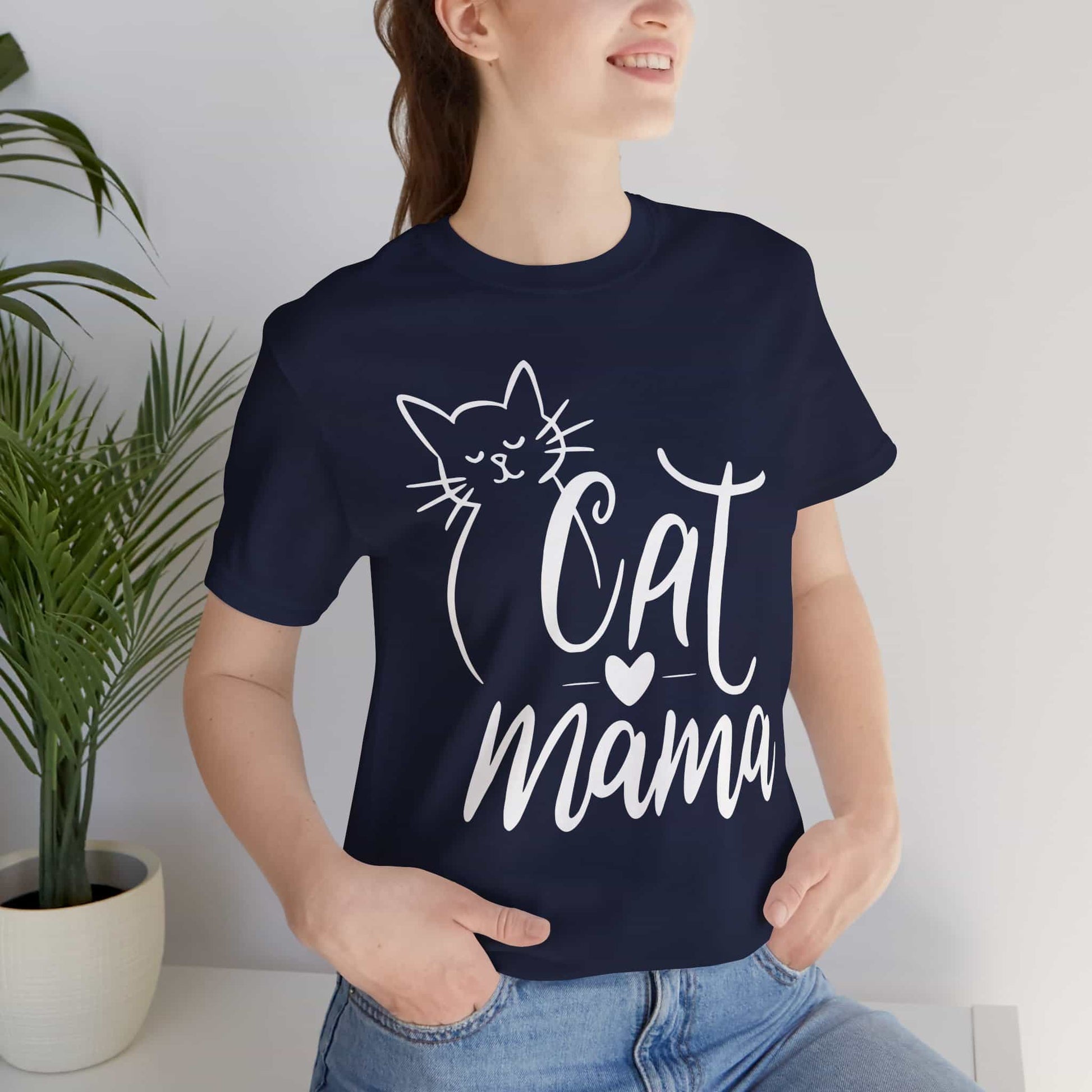 A woman wearing a navy blue Cute Cat Mama T-shirt with 'Cat Mama' design, standing indoors next to a plant.