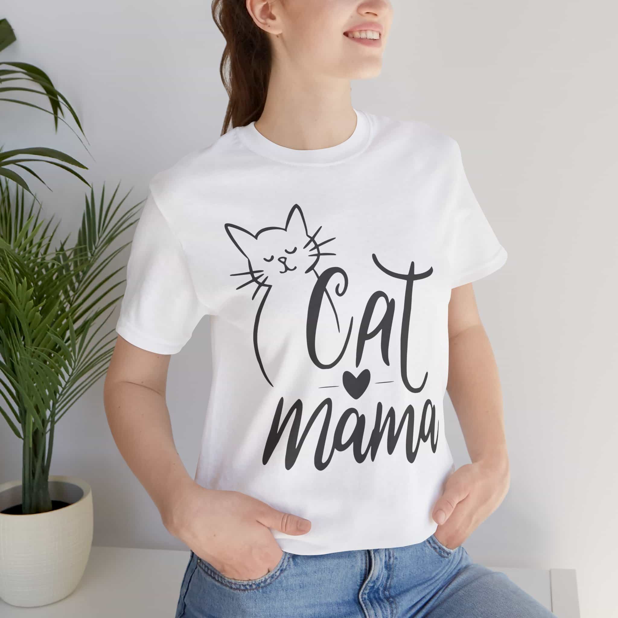 A woman wearing a white Sweet Cat Mama Graphic T-Shirt with 'Cat Mama' design, standing indoors next to a plant.