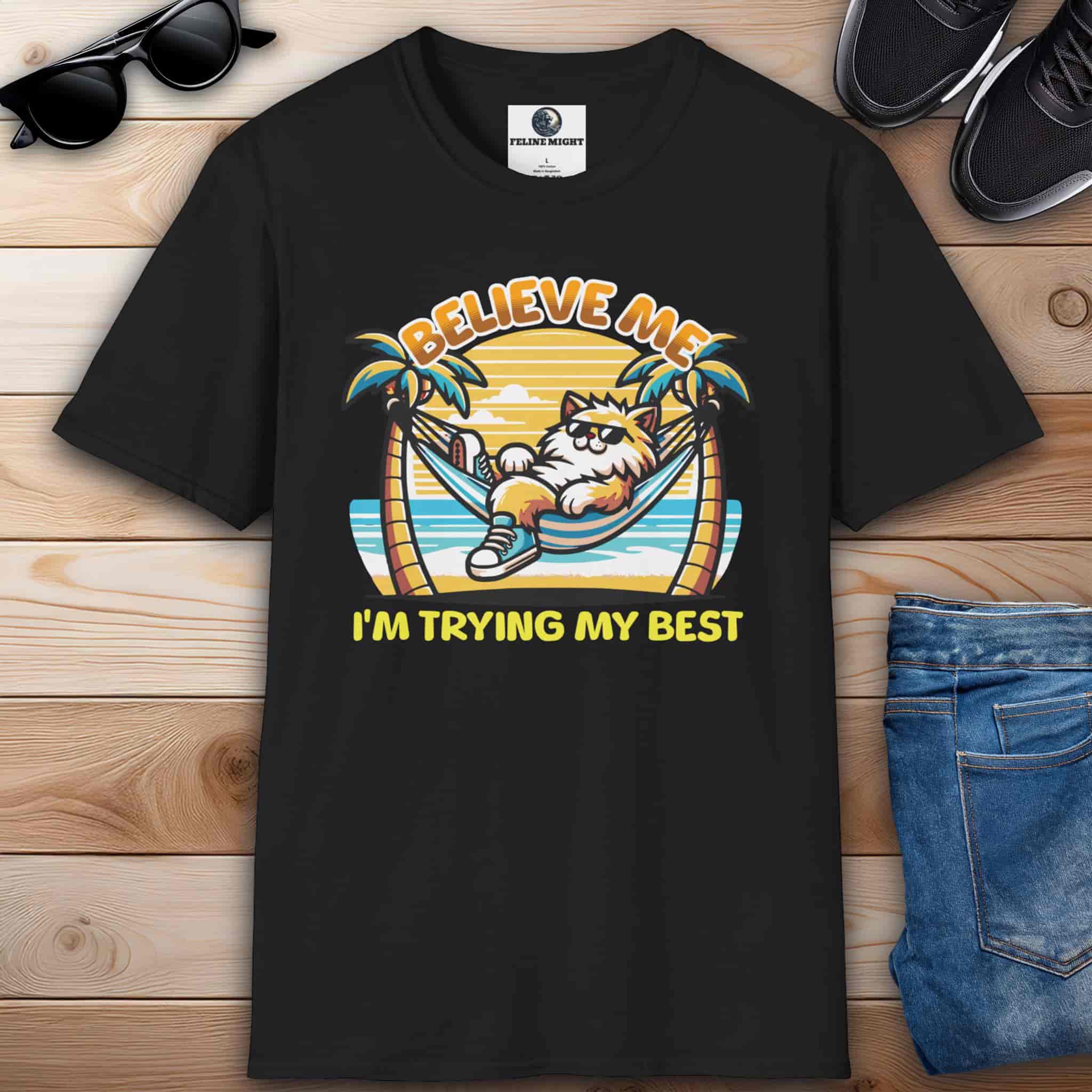 A black t-shirt featuring a cartoon cat relaxing in a hammock with palm trees and the text 'Believe Me I'm Trying My Best'.