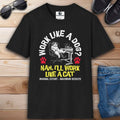 A black Funny Work Cat T-Shirt with a humorous cat graphic and the text 'Work like a dog? Nah, I'll work like a cat,' on a wooden surface with sunglasses and jeans.