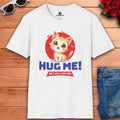 White t-shirt with a cartoon cat and text: "Hug Me! Or I will end you." Surrounded by hearts, laid on a wooden surface next to sunglasses, a red rose bouquet, and blue jeans.