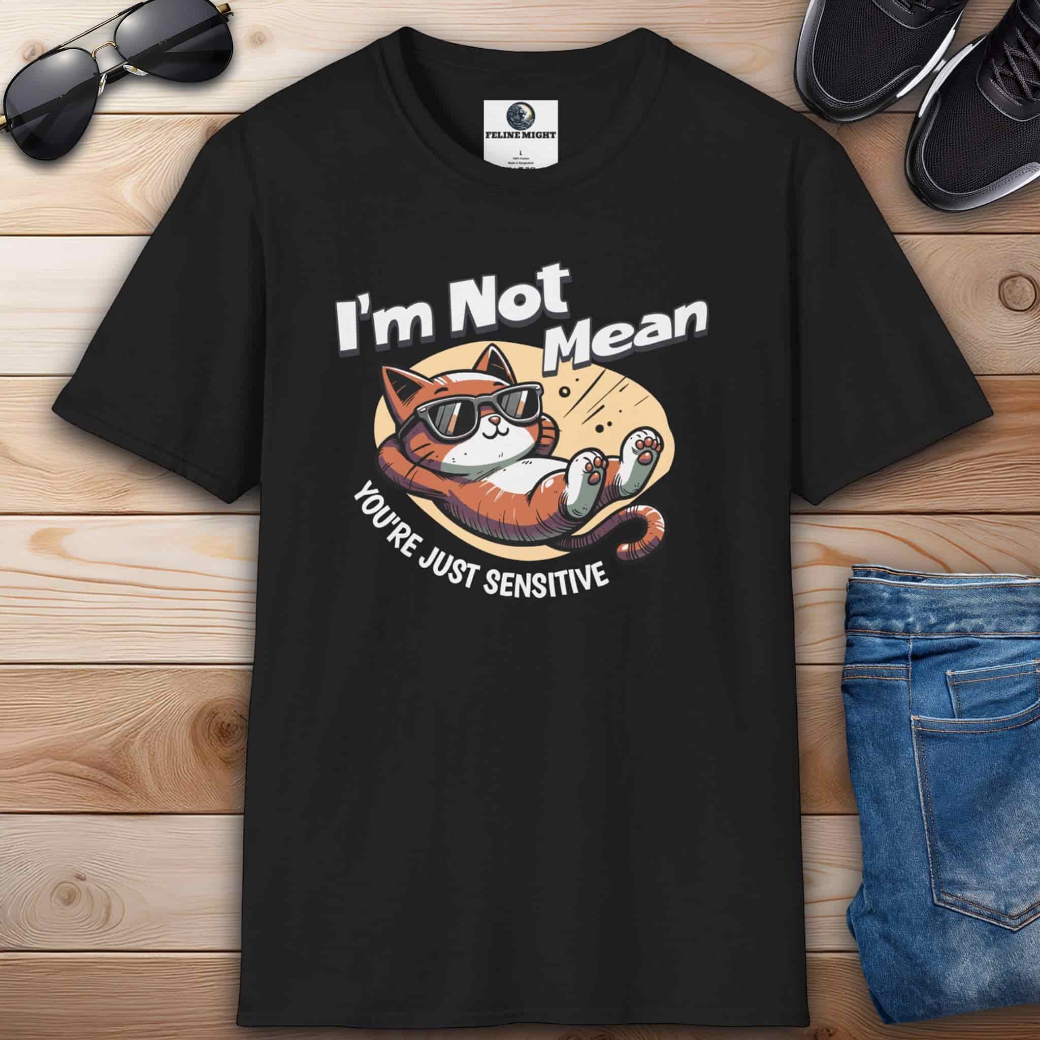 A black Sarcastic Cat Humor T-Shirt with a graphic of a cat wearing sunglasses and the text 'I'm Not Mean, You're Just Sensitive', on a wooden surface with shoes and jeans.