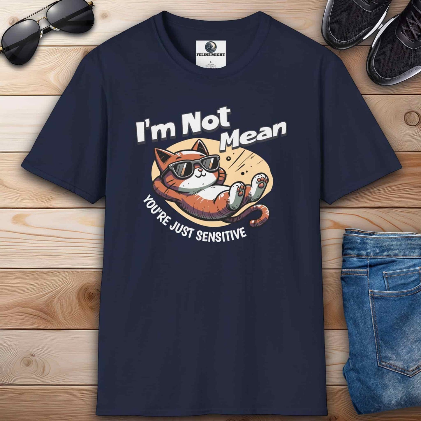 A navy blue Sarcastic Cat Humor T-Shirt with a graphic of a cat wearing sunglasses and the text 'I'm Not Mean, You're Just Sensitive', on a wooden surface with shoes and jeans.