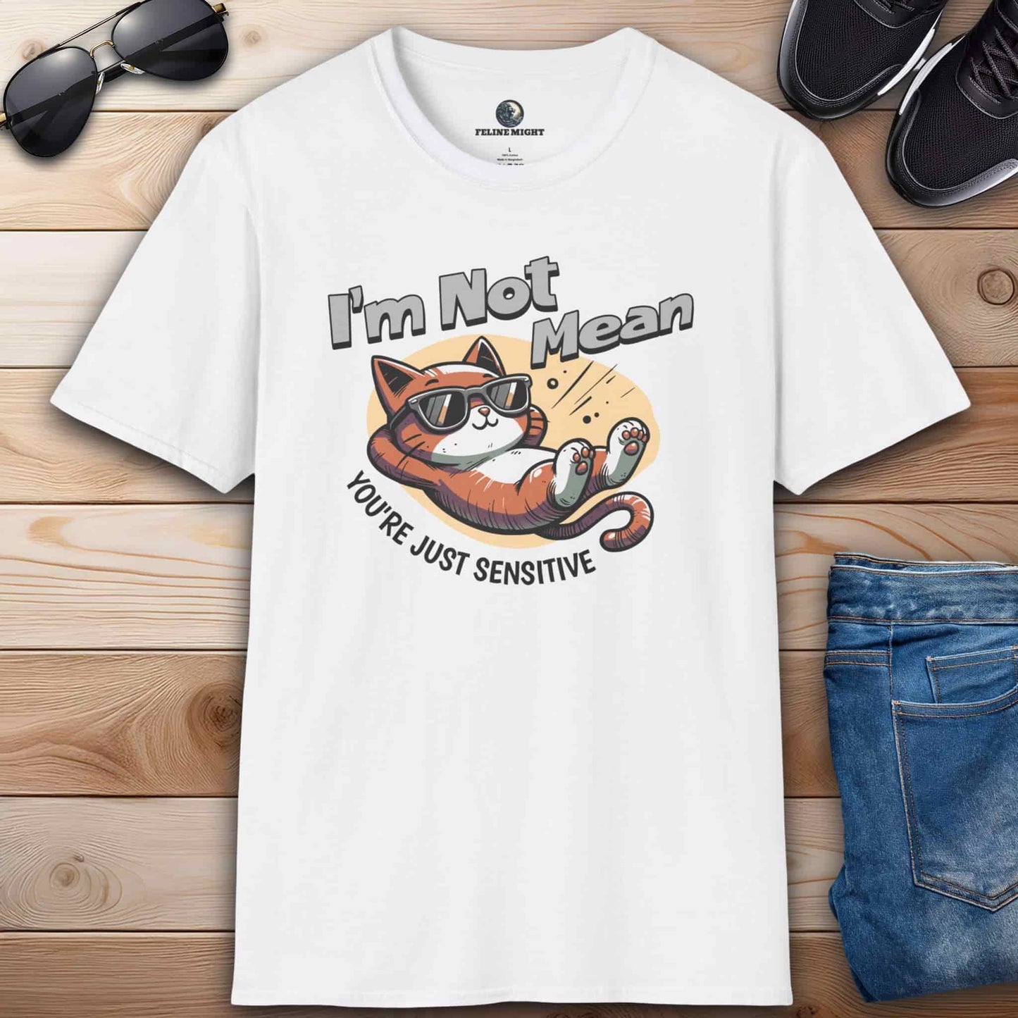 A white Sarcastic Cat Humor T-Shirt with a graphic of a cat wearing sunglasses and the text 'I'm Not Mean, You're Just Sensitive', on a wooden surface with shoes and jeans.