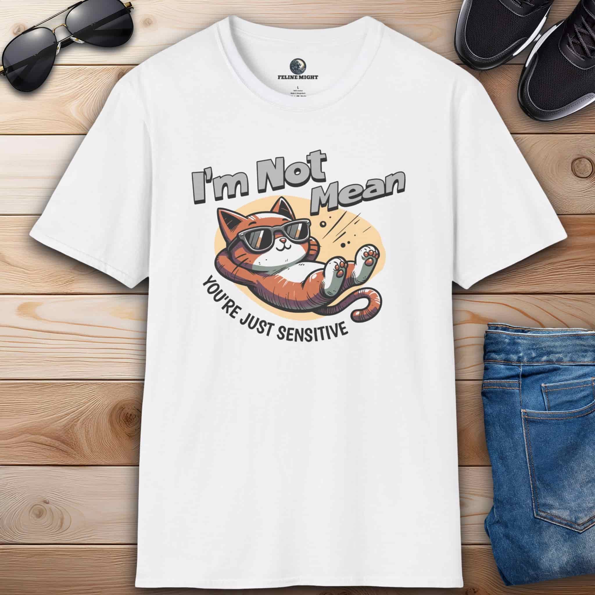 A white Sarcastic Cat Humor T-Shirt with a graphic of a cat wearing sunglasses and the text 'I'm Not Mean, You're Just Sensitive', on a wooden surface with shoes and jeans.